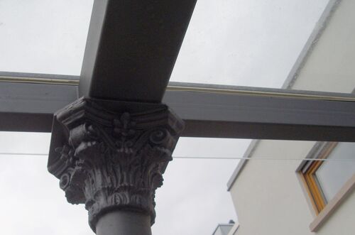 Detailaufnahme einer eleganten Säule unter einer Terrassenüberdachung mit Glasdach und grauem Himmel im Hintergrund.
