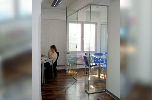 Ein moderner Arbeitsraum mit Glastüren, einem Schreibtisch und einem Besprechungsbereich mit blauen Stühlen.