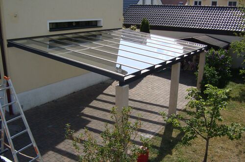 Ein moderner Carport mit Glasdach steht auf einem gepflasterten Parkplatz neben einem Wohnhaus und Garten.