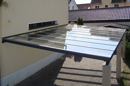 Ein moderner Carport mit transparentem Dach erstreckt sich vor einem Haus auf gepflastertem Boden.