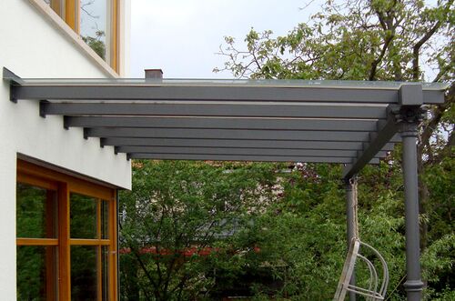 Moderne Terrassenüberdachung mit grauen Balken und eleganten Säulen, umgeben von grünem Laub.